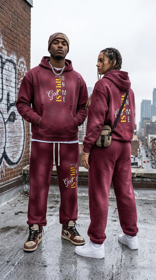 Man and woman wearing matching burgundy God Is Working On Me hoodie and jogger tracksuit set on a city rooftop